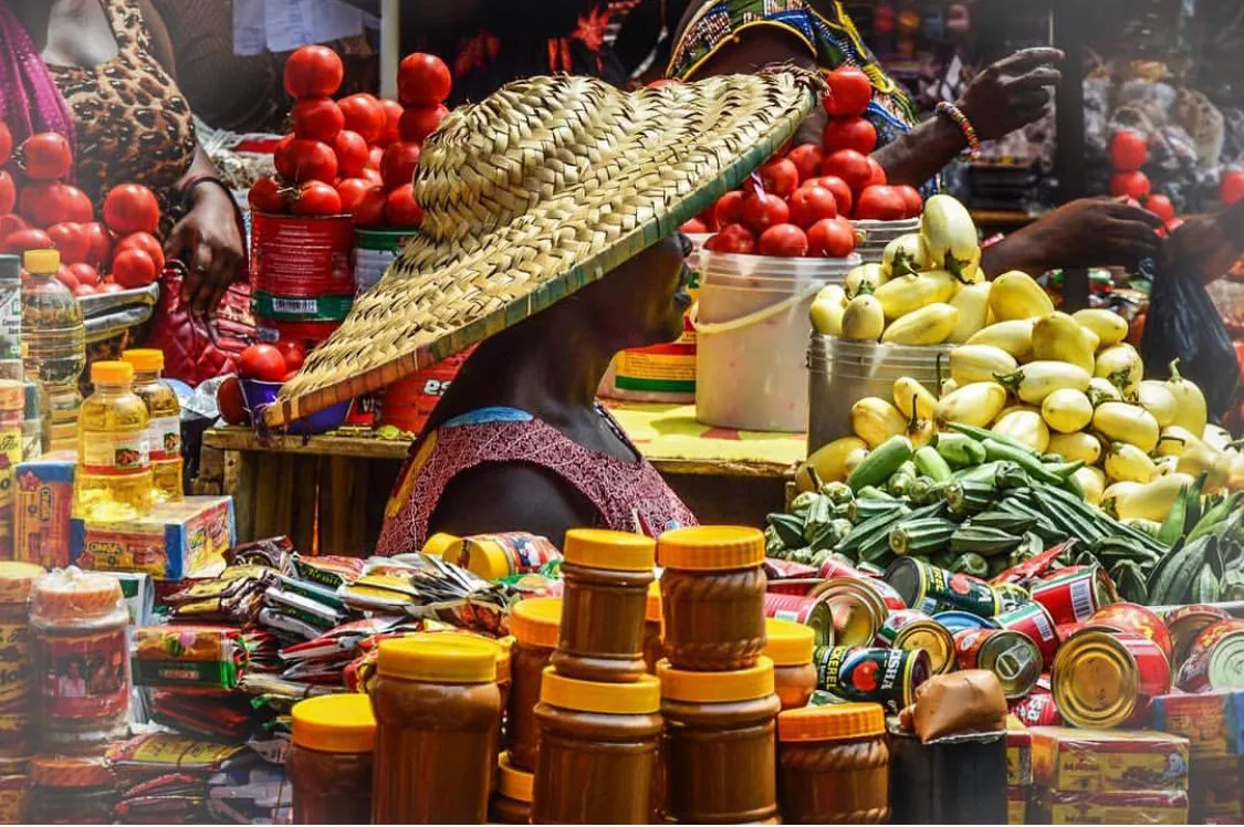 A vendor wearing a large straw hat sells fresh produce, including tomatoes, okra, and bottled goods, in a busy market in Accra, Ghana.